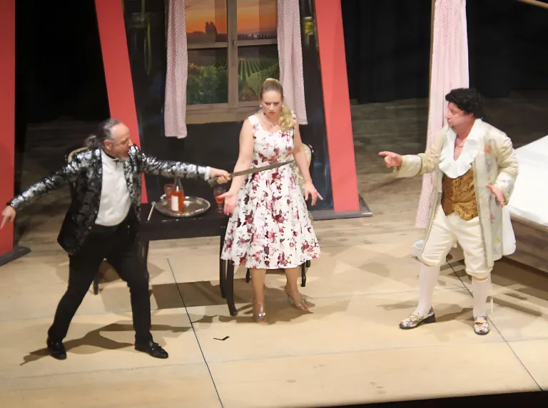 Drei Schauspieler auf der Bühne, zwei Männer und eine Frau, in einer Szene engagiert. Der Mann auf der linken Seite hält ein Schwert, die Frau steht zwischen ihnen. Der Mann auf der rechten Seite trägt ein goldenes Outfit.