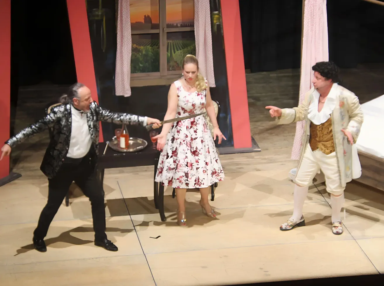 Drei Schauspieler auf der Bühne, zwei Männer und eine Frau, in einer Szene engagiert. Der Mann auf der linken Seite hält ein Schwert, die Frau steht zwischen ihnen. Der Mann auf der rechten Seite trägt ein goldenes Outfit.