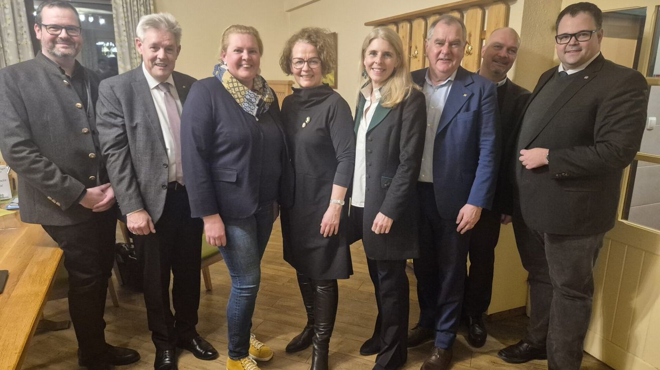 Eine Gruppe von Menschen, sowohl Männer als auch Frauen, steht in einem Raum. Sie sind alle formell gekleidet, einige tragen Krawatten und Lederschuhe. Die Frau in der Mitte trägt eine Brille und einen Schal. Hinter ihnen befindet sich eine Holzwand mit einer gelben Wand.