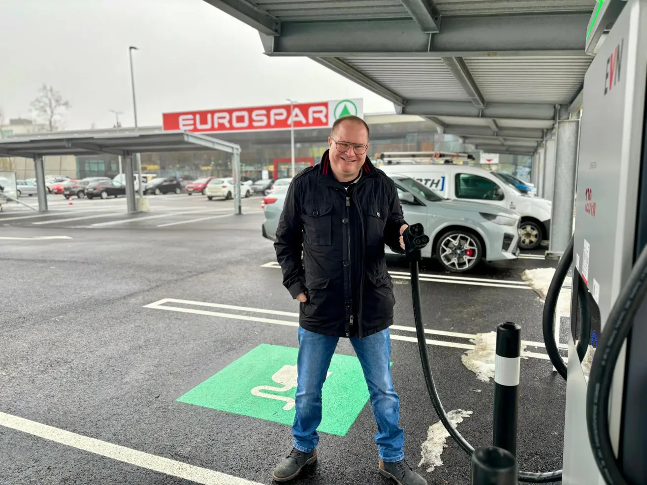 Ein Mann in einer schwarzen Jacke lädt ein Elektroauto an einer Ladestation auf einem Parkplatz auf. Hinter ihm steht ein Gebäude mit einem EuroSpar-Schild.