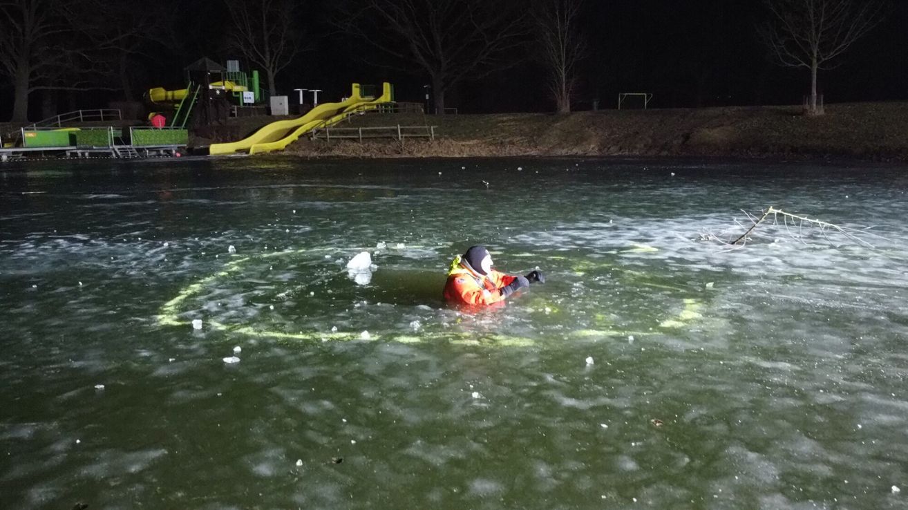 Eine Person steht in einer reflektierenden orangefarbenen Jacke im eisigen Wasser in der Nähe eines gelben Rutschens. Bäume sind sichtbar, und es ist Nacht.