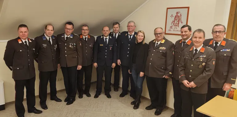 Sieben Personen in Uniform stehen gemeinsam für ein Foto. Sie stehen vor einer Wand mit einem Bilderrahmen. Sie lächeln für die Kamera.