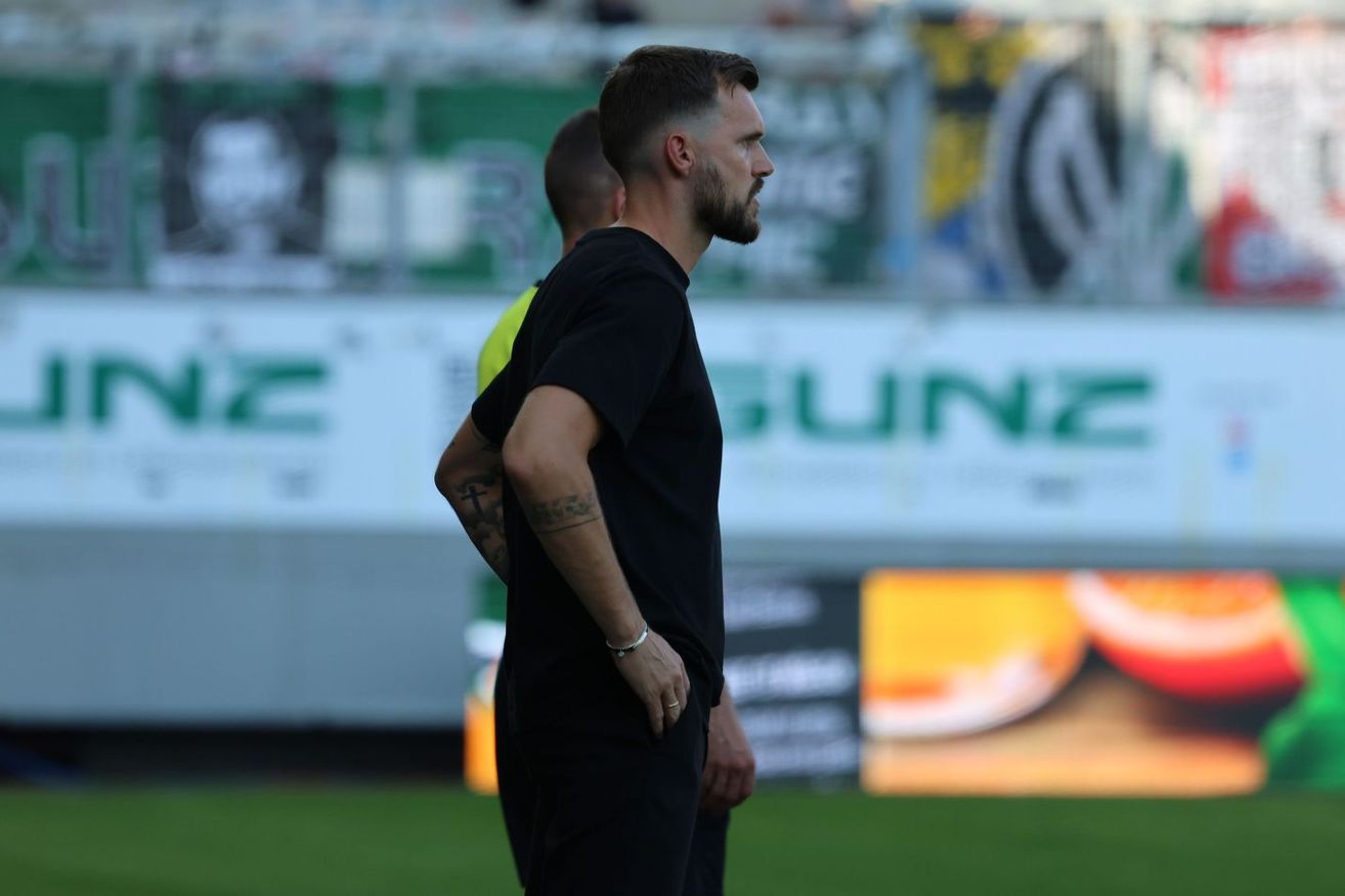 Ein Mann mit Tattoos steht auf einem Fußballfeld, dem Rücken zur Kamera, mit einer weiteren Person hinter ihm. Im Hintergrund befinden sich verschwommene Werbungen und ein Zaun.