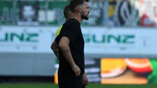 Ein Mann mit Tattoos steht auf einem Fußballfeld, dem Rücken zur Kamera, mit einer weiteren Person hinter ihm. Im Hintergrund befinden sich verschwommene Werbungen und ein Zaun.