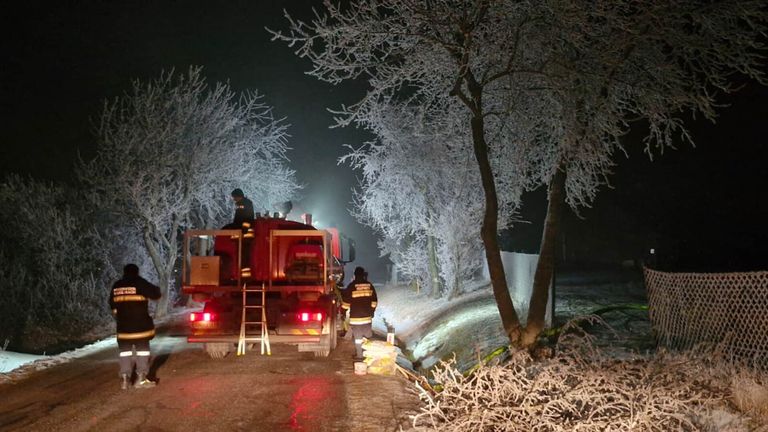 In der Nacht stehen zwei Feuerwehrleute neben einem Feuerwehrwagen auf einer verschneiten Straße, mit vereisten Bäumen und einem Zaun im Hintergrund.
