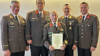 Ein älterer Mann in Uniform steht mit vier anderen Männern. Er hält eine Urkunde in den Händen.