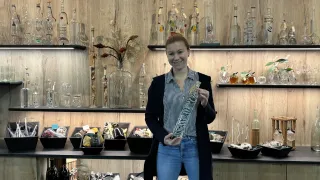 Eine Frau steht in einem Geschäft und hält eine durchsichtige Flasche. Hinter ihr befinden sich Holzregale mit verschiedenen dekorativen Glasflaschen und -gläsern. Auf den Regalen stehen auch schwarze Boxen mit Gegenständen und eine Vase mit Blumen.