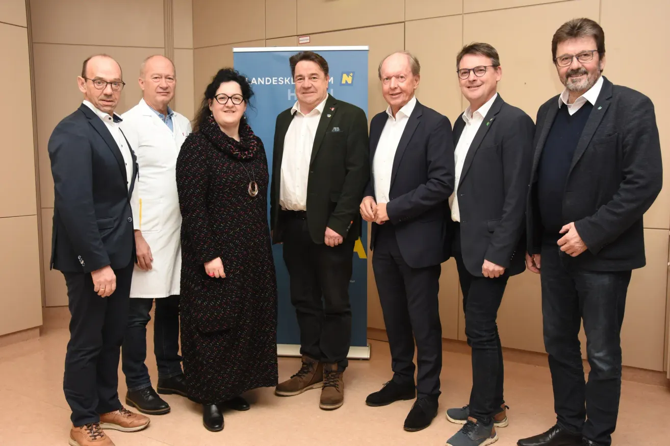 Eine Gruppe von Menschen in formeller Kleidung, einige mit Brille, steht gemeinsam vor einem Banner. Eine Frau mit Brille und Halskette steht zwischen zwei Männern in Anzügen. Dahinter ist ein Banner mit blauem Text und einem Logo zu sehen.