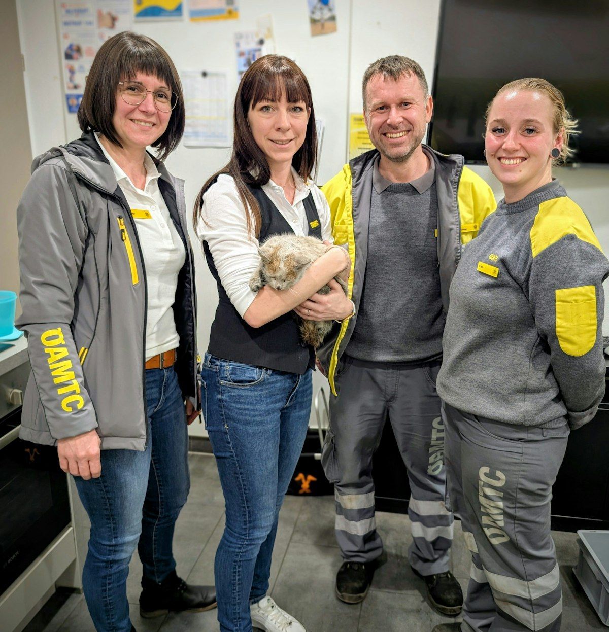 Vier Personen stehen in einem Raum mit einer grau-gelben Jacke, eine von ihnen hält ein kleines Tier. Sie alle haben fröhliche Gesichtsausdrücke.