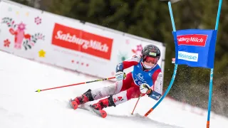 Ein Skifahrer in roter und weißer Ausrüstung fährt einen verschneiten Hang hinunter, wobei Schnee aufspritzt. Dahinter steht eine Wand mit 'Saalburg-Milch' und 'Saalburg' Bannern.