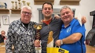 Drei Männer stehen lächelnd nebeneinander, einer hält einen goldenen Pokal und der andere einen gelben Beutel. Der Mann in der Mitte trägt einen grauen Anzug.