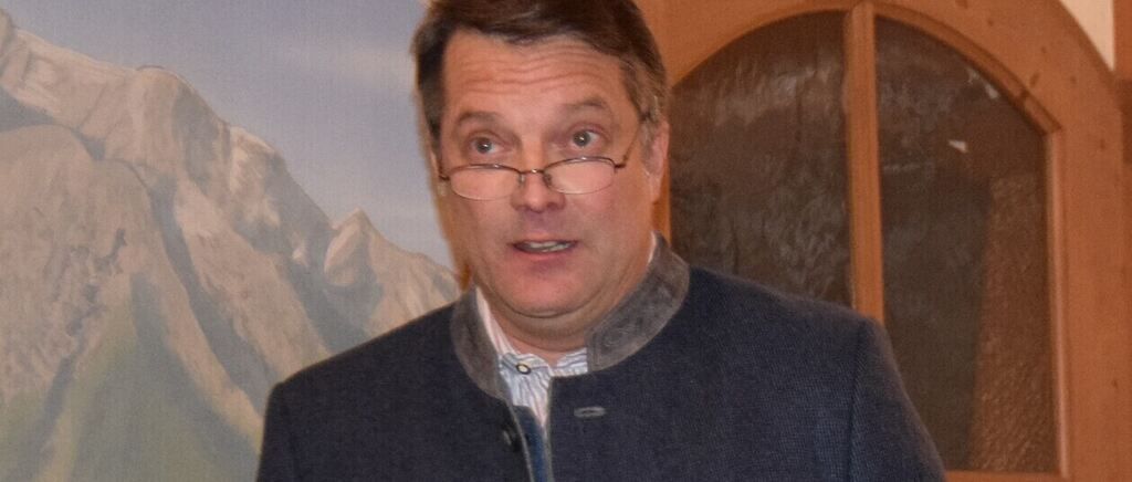 Ein Mann mit Brille und blauem Pullover steht vor einer Wand mit einem Fenster und einem Berggemälde.