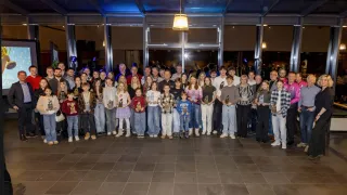Eine große Gruppe von Kindern und Erwachsenen posiert für ein Foto in einem Saal. Sie halten Auszeichnungen und einige tragen Jacken. Der Raum ist gut beleuchtet, mit einer großen Tür und Glaswänden.
