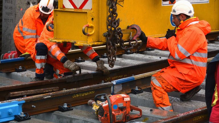 Arbeiter in orangefarbenen Overalls und Helmen benutzen Werkzeuge und Ketten, um ein gelbes Objekt, vermutlich ein Gleis, anzuheben, während sie auf Betonblöcken stehen. Ein roter Kettensäge liegt in der Nähe.