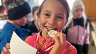 Ein junges Mädchen hält eine goldene Medaille im Mund, trägt eine rote Jacke und steht unter anderen Kindern.