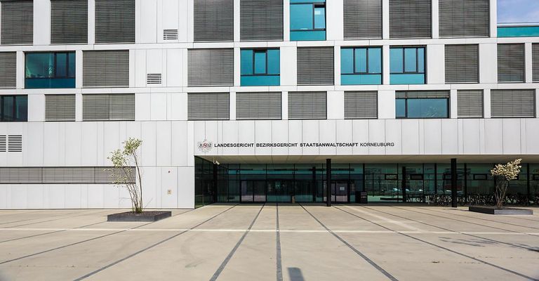 Ein modernes Gebäude mit weißen Wänden, Glasfenstern und einem Schild mit der Aufschrift Landesgericht Bezirksgericht Staatsanwaltsschaft Korneuburg. Links befindet sich eine Pflanze in einem Pflanzgefäß.