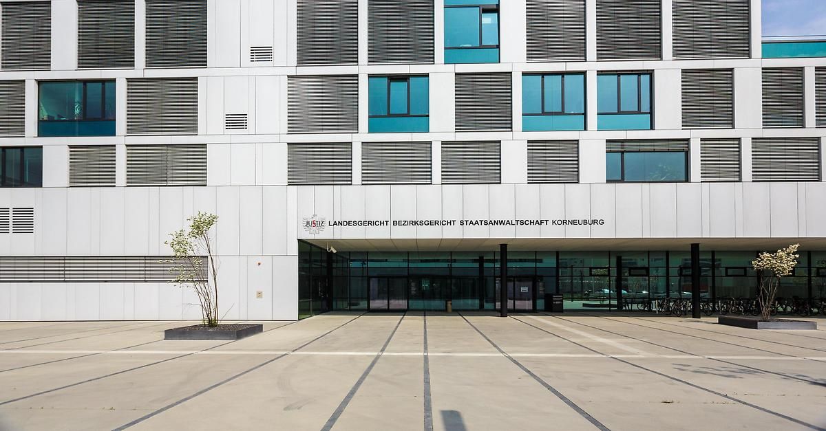 Ein modernes Gebäude mit weißen Wänden, Glasfenstern und einem Schild mit der Aufschrift Landesgericht Bezirksgericht Staatsanwaltsschaft Korneuburg. Links befindet sich eine Pflanze in einem Pflanzgefäß.