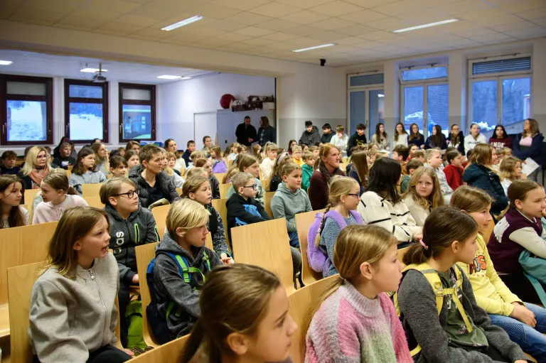 Eine große Gruppe von Schülern in einem Klassenzimmer. Viele tragen Winterkleidung und Rucksäcke. Einige stehen in der Nähe des Eingangs, während andere in Reihen sitzen. Der Raum hat Fenster und Lampen an der Decke.