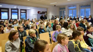 Eine große Gruppe von Schülern in einem Klassenzimmer. Viele tragen Winterkleidung und Rucksäcke. Einige stehen in der Nähe des Eingangs, während andere in Reihen sitzen. Der Raum hat Fenster und Lampen an der Decke.