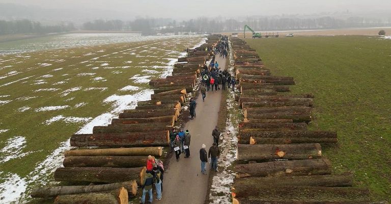 Eine Gruppe von Menschen geht auf einem von Baumstämmen gesäumten Pfad in einer verschneiten Landschaft. In der Ferne sind ein Kran und Fahrzeuge zu sehen.