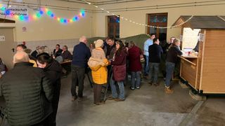 Eine Gruppe von Menschen versammelt sich in einem Raum, einige stehen in der Nähe eines Tisches, andere unterhalten sich. Bunte Lichter schmücken die Wände, und im Hintergrund ist ein Zelt aufgebaut.