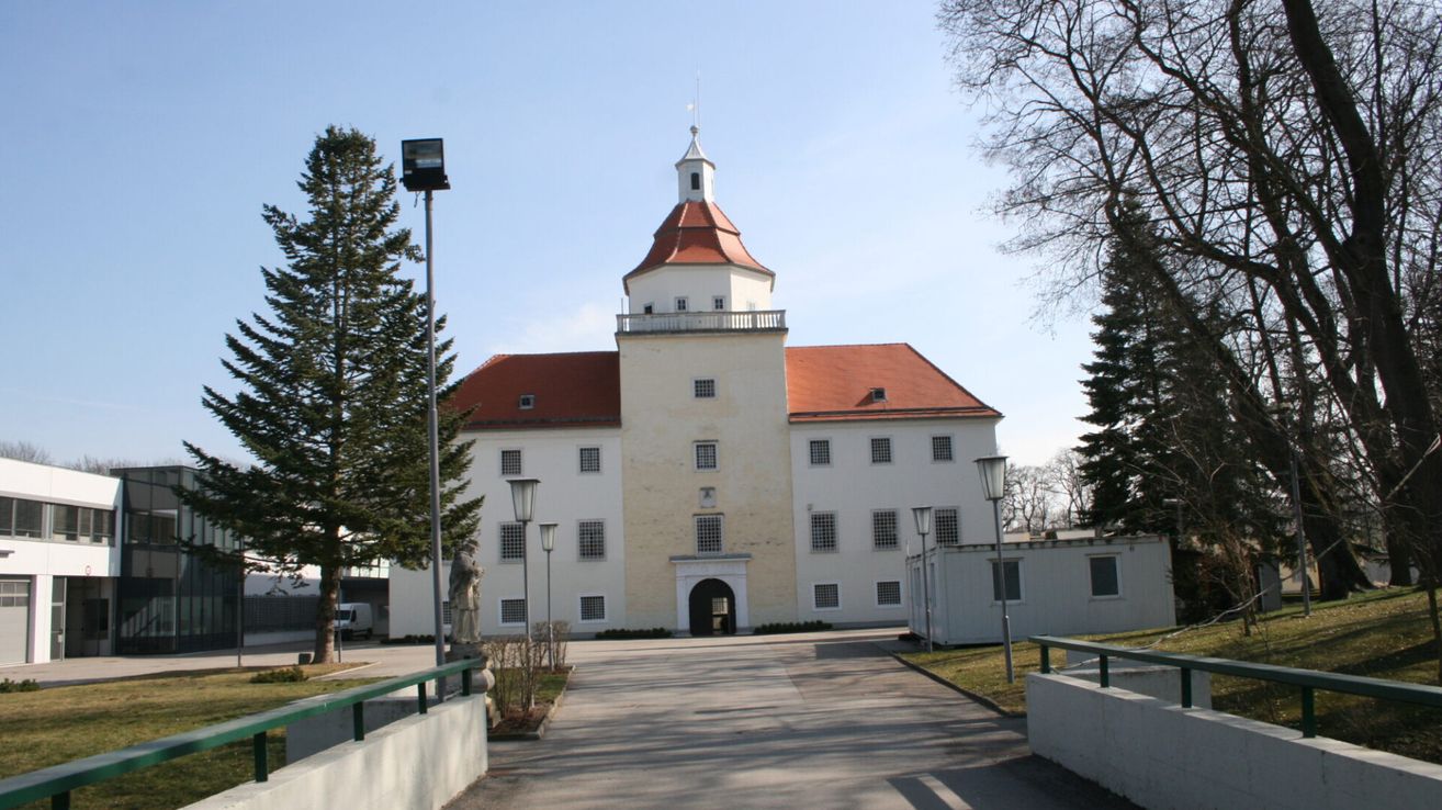 Ein großes, weißes Gebäude mit rotem Dach und Turm. Der Eingang wird von zwei Straßenlampen flankiert. Links steht eine Statue, und in der Nähe befinden sich Bäume und Büsche.