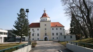 Ein großes, weißes Gebäude mit rotem Dach und Turm. Der Eingang wird von zwei Straßenlampen flankiert. Links steht eine Statue, und in der Nähe befinden sich Bäume und Büsche.