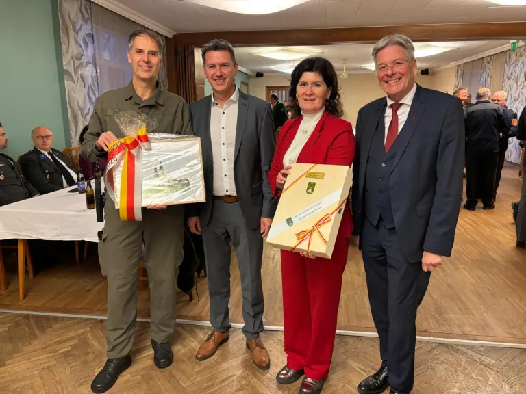 Vier Menschen stehen lächelnd zusammen in einem Raum. Zwei Männer und zwei Frauen. Eine Frau hält eine Geschenkbox, eine andere ein gerahmtes Bild. Sie tragen formelle Kleidung. Ein Tisch mit Flaschen und einem Ventilator im Hintergrund.