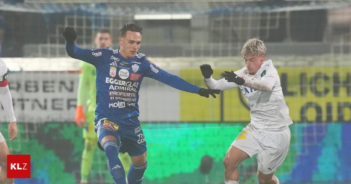 Zwei Fußballer im Einsatz, einer in Blau mit 'EGGERGLAS' und 'PROFERTIL' auf seinem Trikot, der andere in Weiß, beide mit Handschuhen. Sie ringen eng um den Ball. Im Hintergrund ist ein verschwommener Hintergrund mit einem grün-gelben Banner.