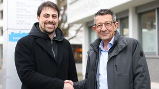 Zwei Männer in Jacken schütteln sich die Hände draußen. Ein Mann lächelt, der andere trägt eine Brille. Hinter ihnen steht ein Gebäude mit Glasfenstern.