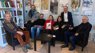 Sieben Personen, darunter Männer und Frauen, sitzen in einem Raum, halten Bücher in der Hand und lächeln. Der Raum hat ein weißes Bücherregal voller Bücher, einen gefliesten Boden und mehrere Poster an der Wand.