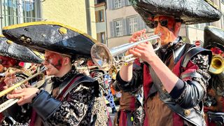 Eine Gruppe von Menschen in Piratenkostümen spielt Blechblasinstrumente auf der Straße, einer spielt Trompete, der andere Posaune, mit einem Gebäude im Hintergrund.