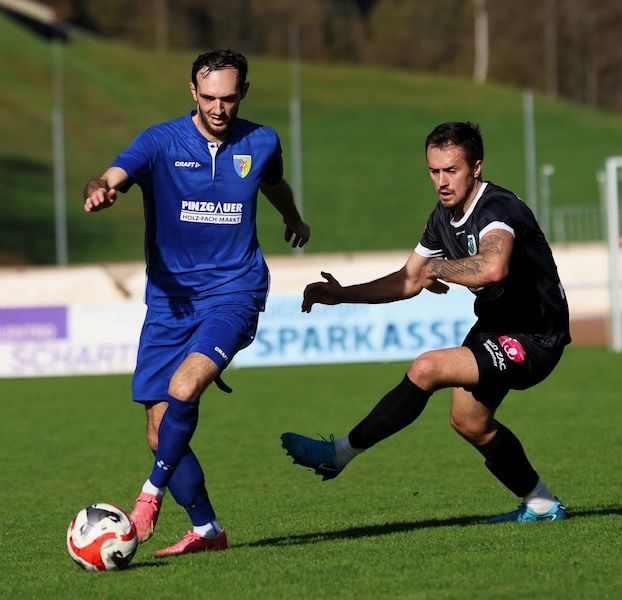 Zwei Fußballspieler, einer in Blau und einer in Schwarz, konkurrieren auf einem Feld mit einem Fußball vor ihnen. Der Spieler in Blau hat ein Sponsorenlogo auf seinem Trikot.