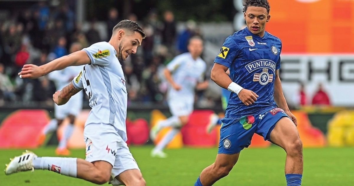 Zwei Fußballspieler auf einem Spielfeld, einer in Weiß und einer in Blau, laufen mit einem verschwommenen Hintergrund von Zuschauern.