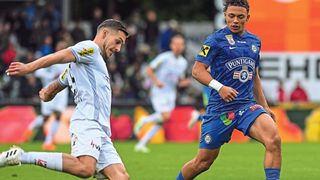 Zwei Fußballspieler auf einem Spielfeld, einer in Weiß und einer in Blau, laufen mit einem verschwommenen Hintergrund von Zuschauern.