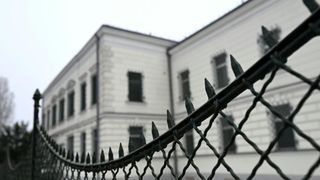 Ein weißes Gebäude mit mehreren Fenstern und davor ein Maschendrahtzaun mit spitzen Enden.