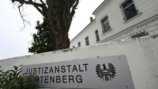 Ein Schild mit dem Wort 'SITZANSTALT' und dem österreichischen Adlerwappen. Es ist an einer weißen Wand befestigt, und dahinter befindet sich ein Gebäude mit einem Schornstein und mehreren Fenstern.