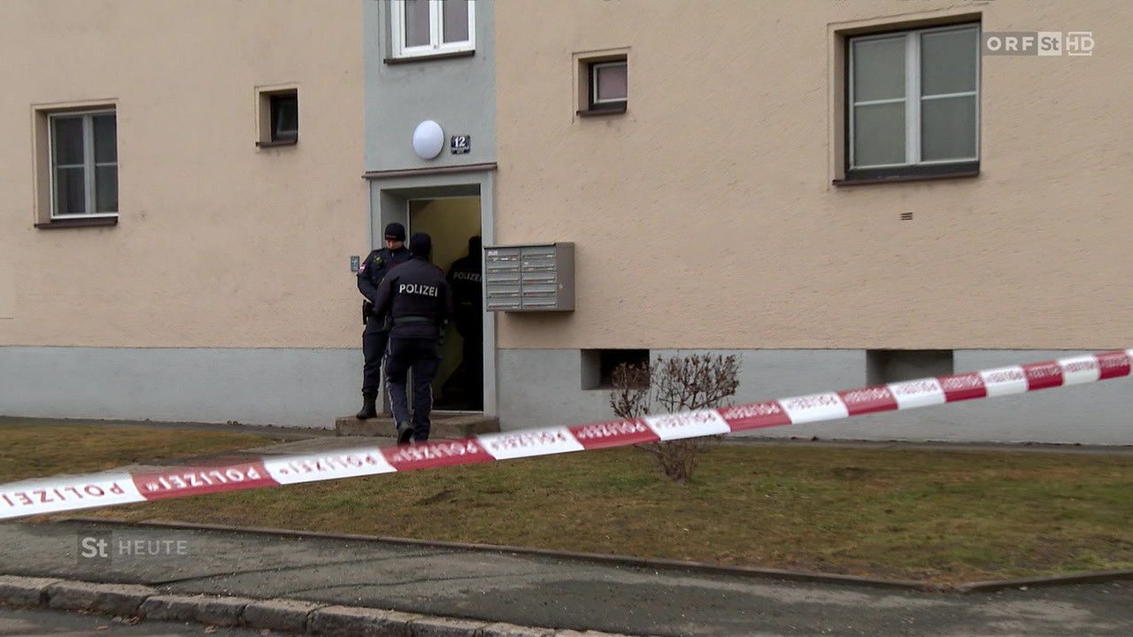 Zwei Polizisten stehen vor einem Gebäude, wahrscheinlich im Rahmen einer Ermittlung. Der Eingang ist mit einem Band abgesperrt, und ein Briefkasten ist an der Wand montiert.