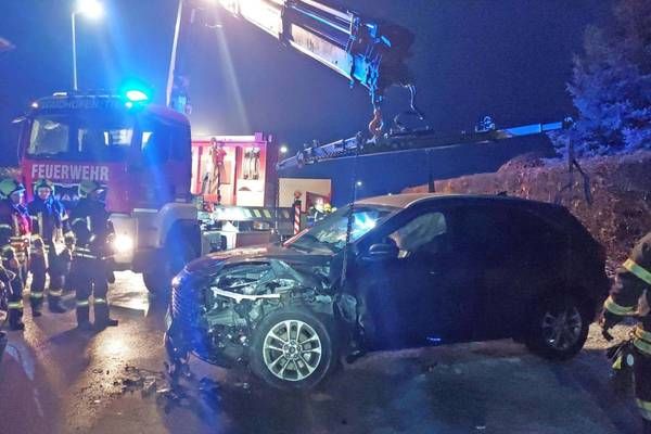 Ein beschädigtes Auto wird nachts von einem Kran angehoben, während Feuerwehrleute darum stehen.