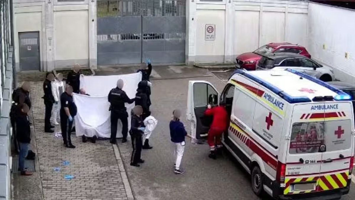 Rettungskräfte handhaben ein weißes Laken, das einen Körper bedeckt, in der Nähe eines Krankenwagens. Einige Personen in Schutzausrüstung stehen hinter dem Laken. Ein Van ist geparkt mit offener Tür.