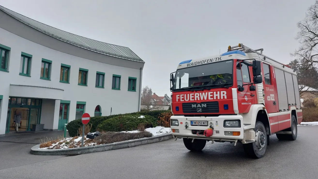 Ein roter und weißer Feuerwehrwagen mit der Aufschrift 'Feuerwehr' fährt an einem Gebäude mit grünen Fensterrahmen und einem rot-weißen Schild vorbei.