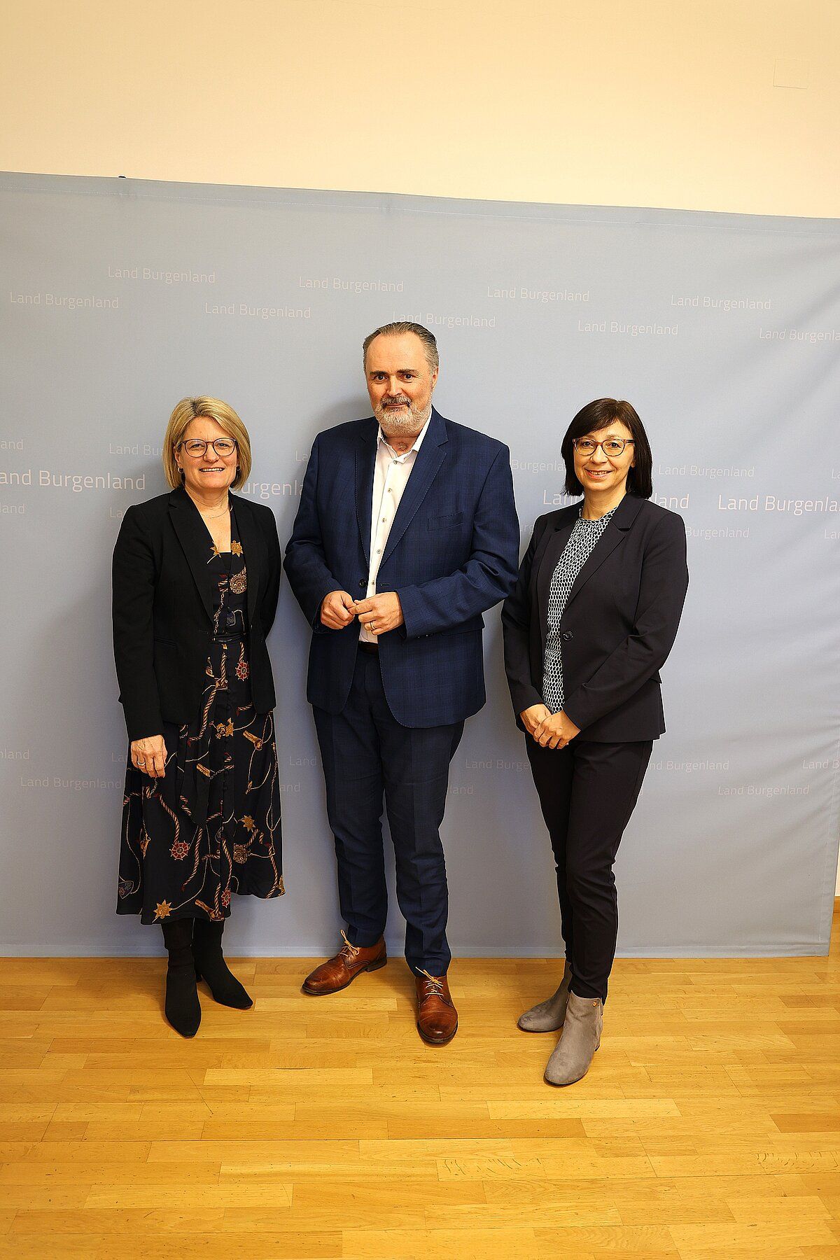 Drei Personen stehen zusammen für ein Foto, möglicherweise für eine Pressekonferenz. Sie alle lächeln. Der Mann trägt einen Anzug und die Frauen formelle Kleidung. Hinter ihnen befindet sich ein Hintergrund mit dem Text 'Land Burgenland'.