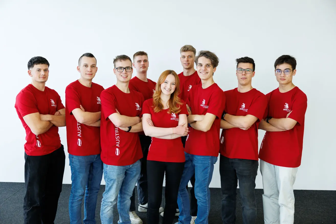 Eine Gruppe von Menschen in roten T-Shirts posiert für ein Foto mit einer lächelnden Frau in der Mitte. Sie stehen mit verschränkten Armen vor einer weißen Wand.