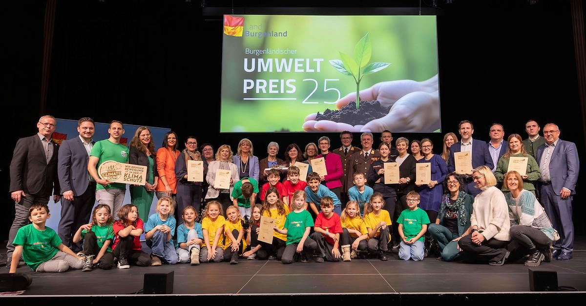 Eine Gruppe von Kindern und Erwachsenen posiert für ein Foto auf einer Bühne mit einem Banner und Zertifikaten. Ein großer Bildschirm hinter ihnen zeigt eine Hand, die eine Pflanze hält.