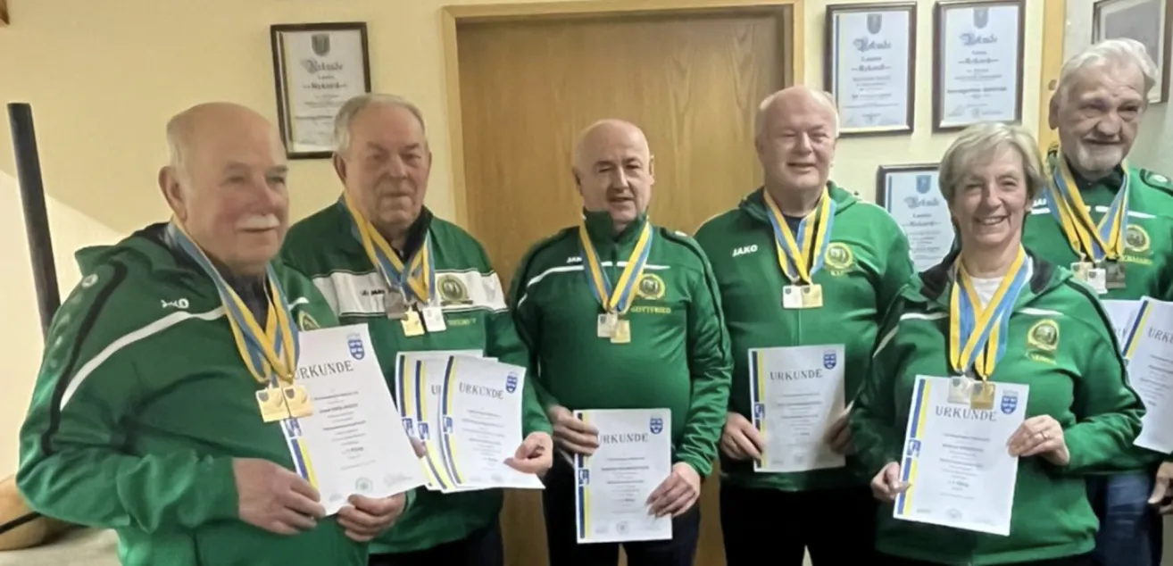 Vier Männer in grünen Jacken und Medaillen posieren für ein Foto. Sie halten Urkunden mit dem Wort Urkunde. Hinter ihnen ist eine Holztür mit einem Schild.