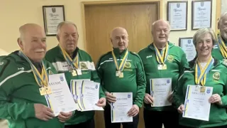 Vier Männer in grünen Jacken und Medaillen posieren für ein Foto. Sie halten Urkunden mit dem Wort Urkunde. Hinter ihnen ist eine Holztür mit einem Schild.