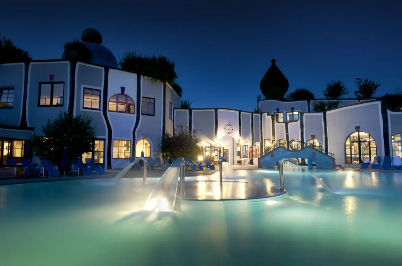 Ein modernes architektonisches Resort mit einem Pool bei Nacht. Das Gebäude hat eine Uhr, mehrere Fenster und ist von Liegestühlen umgeben. Wasserfontänen sind im Pool beleuchtet.