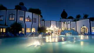 Ein modernes architektonisches Resort mit einem Pool bei Nacht. Das Gebäude hat eine Uhr, mehrere Fenster und ist von Liegestühlen umgeben. Wasserfontänen sind im Pool beleuchtet.