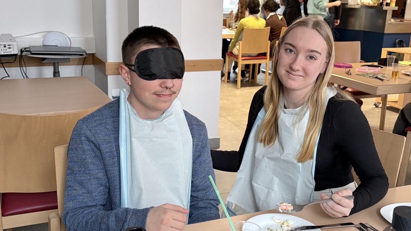 Ein junger Mann mit einem Augenverband sitzt am Tisch und hält eine Gabel. Neben ihm steht eine lächelnde Frau mit einem Schürzen, beide tragen Schürzen. Im Hintergrund sitzen andere Personen an Tischen.