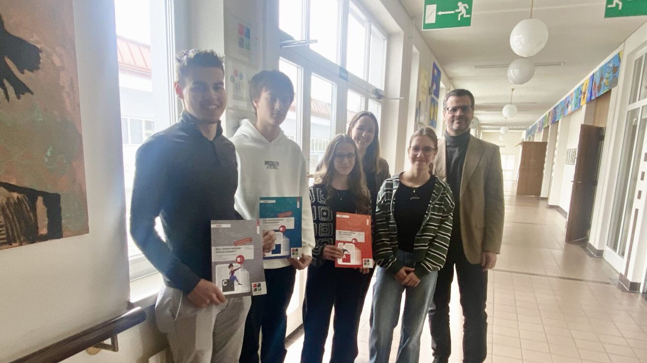 Fünf Personen stehen in einem Flur und halten Bücher in den Händen. Sie lächeln für ein Foto. Zwei Männer tragen Freizeitkleidung, einer in Schwarz, der andere in Weiß. Drei Frauen, zwei in Schwarz und eine in einer gestreiften Jacke, halten Bücher und lächeln. Der Flur hat Glasfenster und ein Ausfahrtsschild.